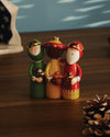 Colorful Three Wise Men ceramic figurines by Alessi, each holding a golden gift, on a wooden table next to a pinecone.