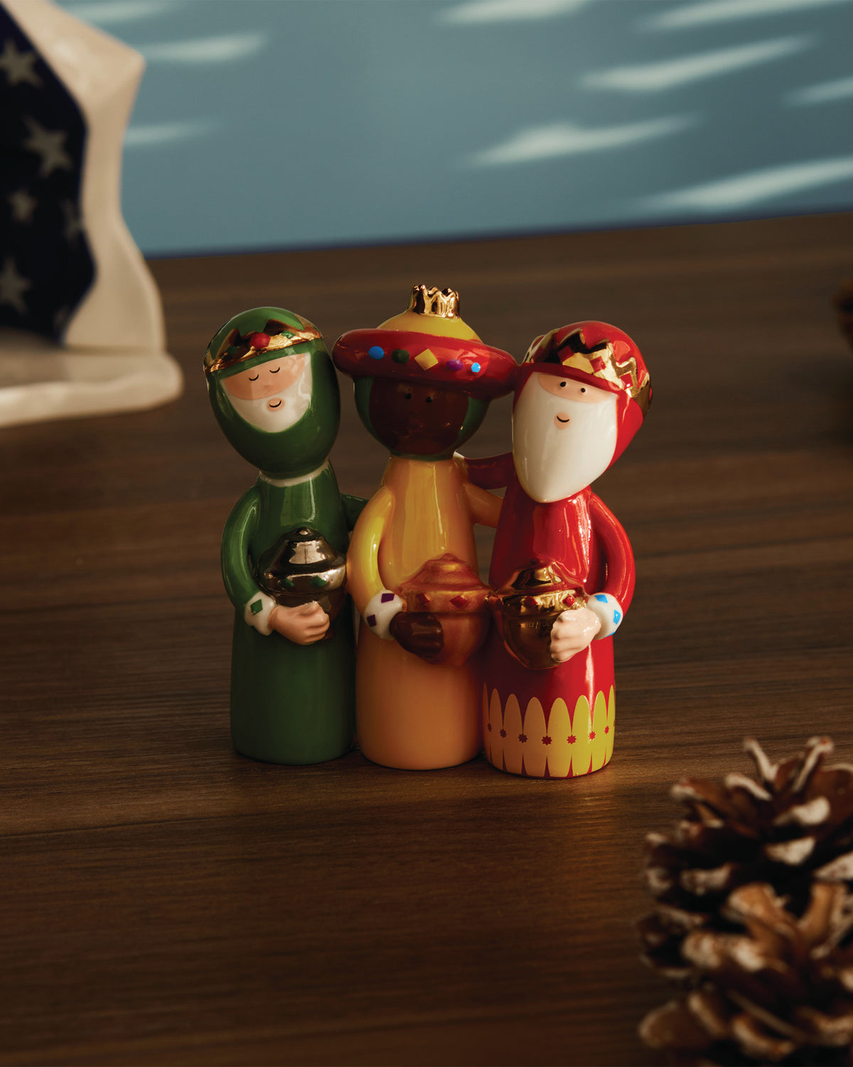Colorful Three Wise Men ceramic figurines by Alessi, each holding a golden gift, on a wooden table next to a pinecone.