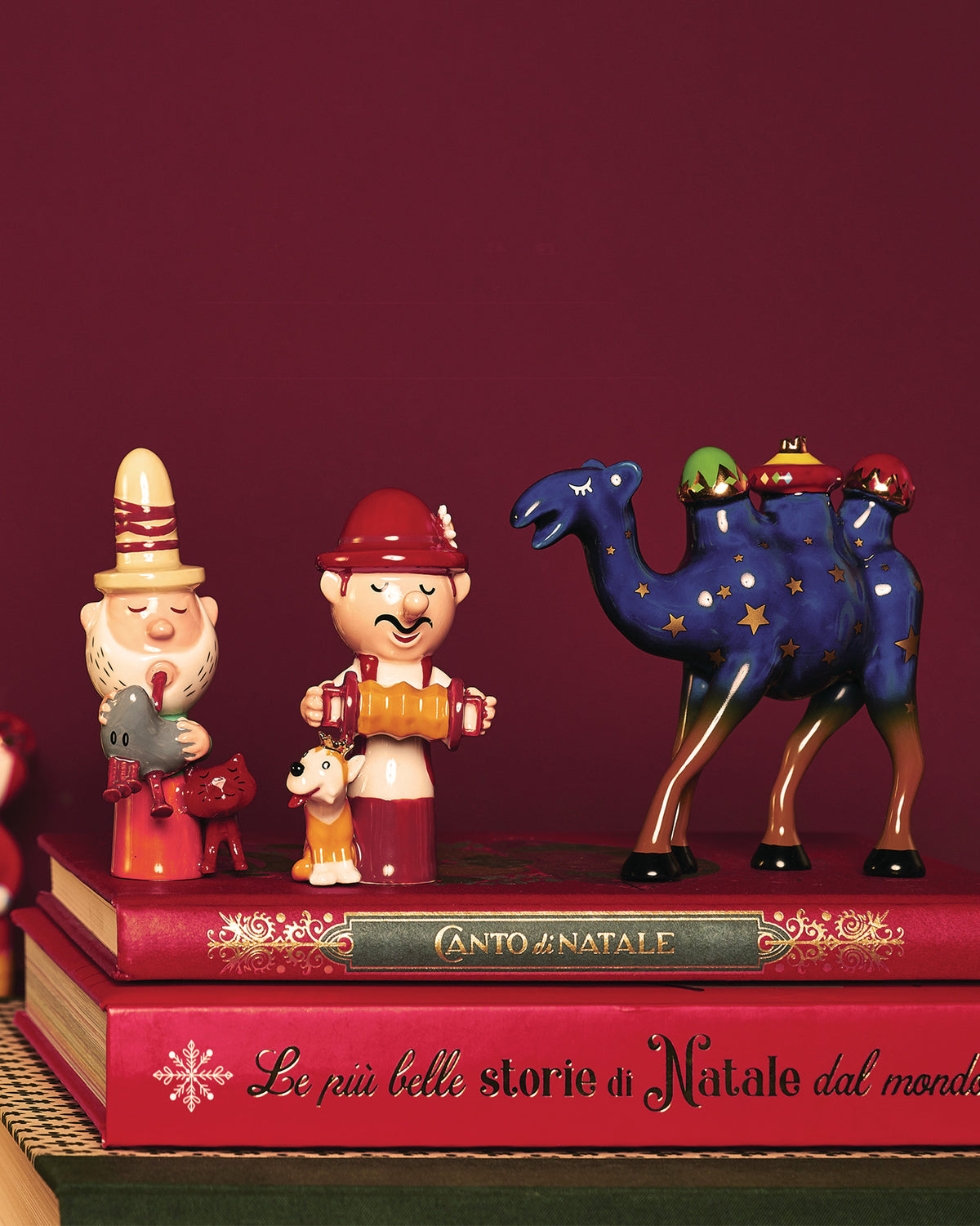 Red Elisabetto ceramic figurine by Alessi, a bearded man in a tall striped hat holding a bird, displayed with other Christmas figures on books.