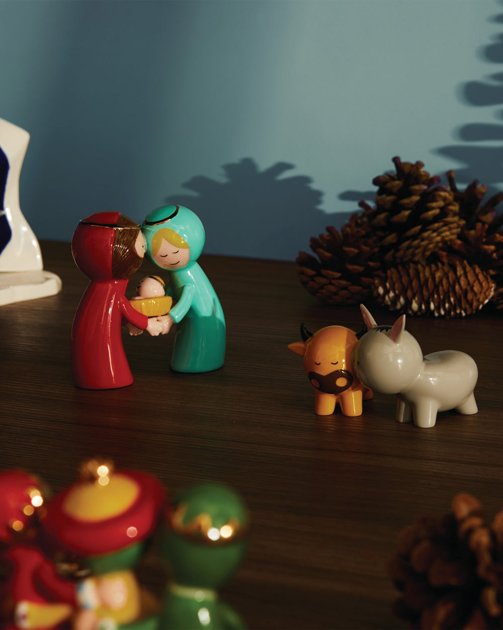 Modern Happy Eternity Baby nativity set by Alessi, with colorful ceramic figures of Mary, Joseph, baby Jesus, an ox, and a donkey, on a wood table with pinecones.