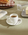 White ceramic espresso cup by Alessi on a saucer with a spoon, part of a sunny breakfast spread.