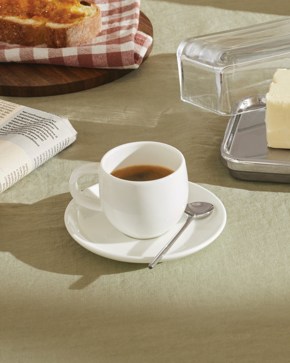 White ceramic espresso cup by Alessi on a saucer with a spoon, part of a sunny breakfast spread.