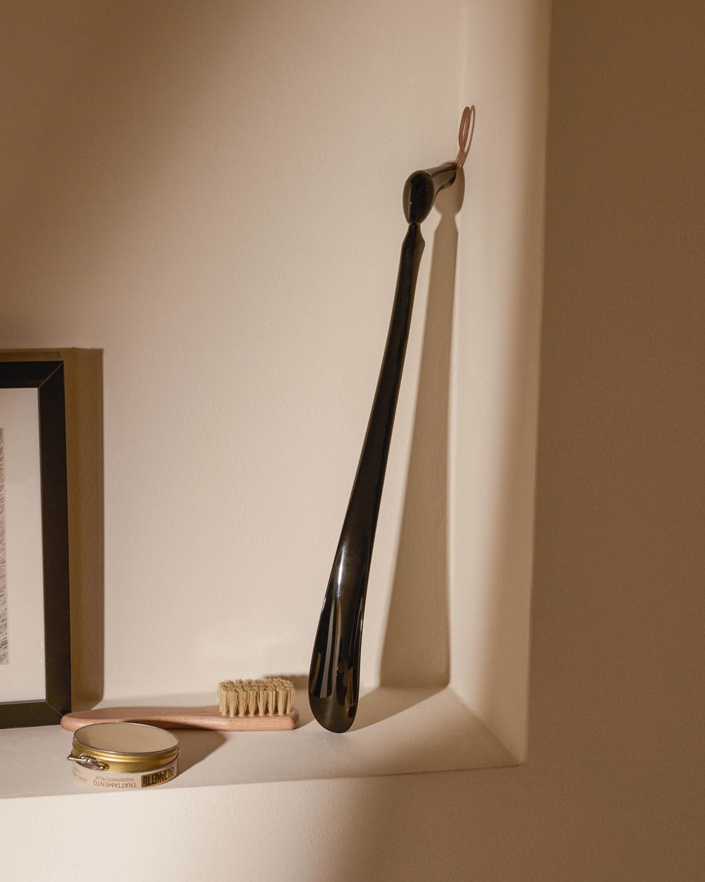 Black glossy shoehorn by Alessi with a brown leather loop, hanging on a wall above shoe polish and a brush.