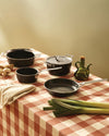Black non-stick pots and pans set by Alessi, with silver handles and lids, on a red and white checkered table.