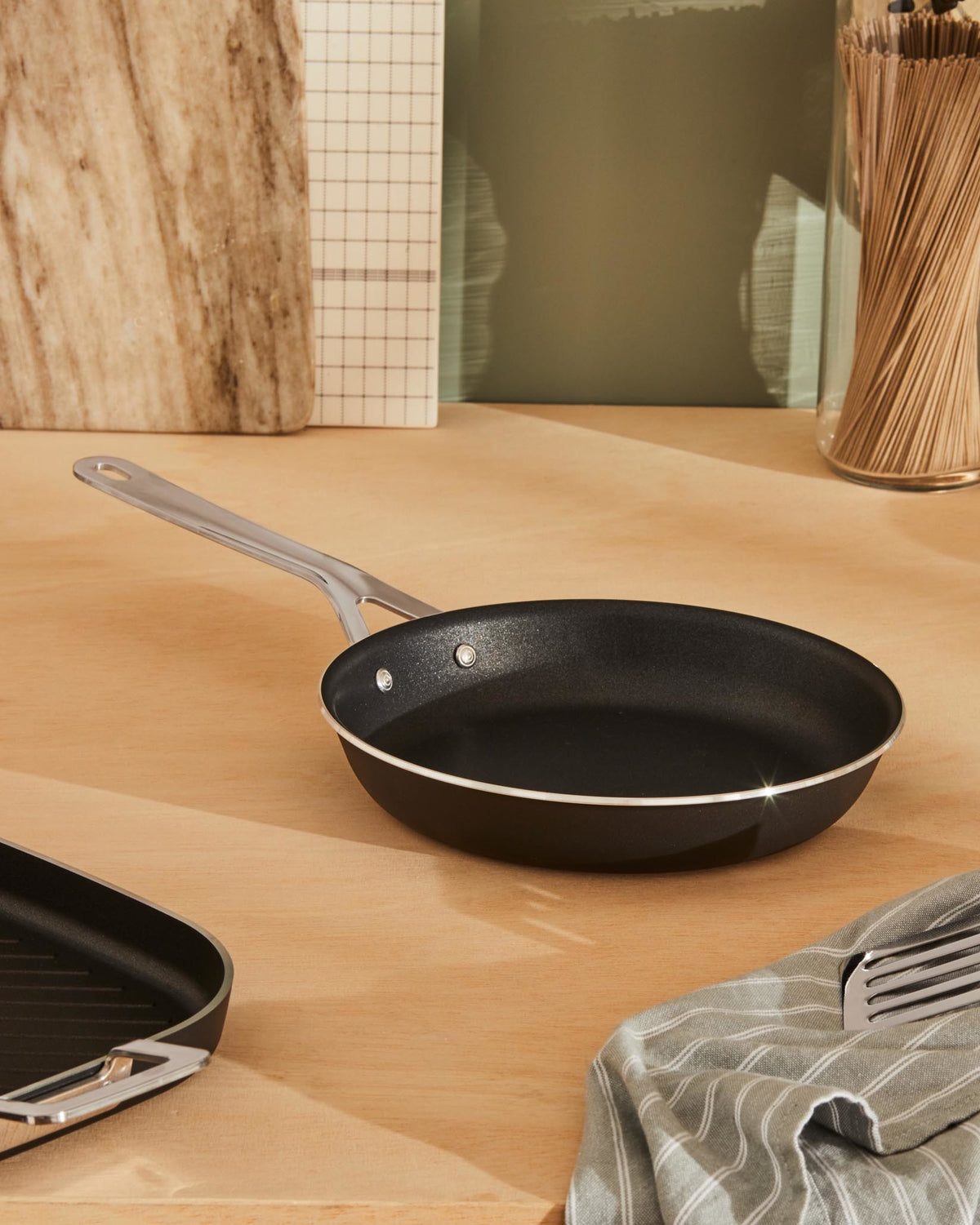 Black non-stick frying pan by Alessi with a silver handle, resting on a wooden counter beside a striped towel.