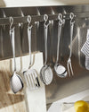 Polished stainless steel kitchen utensil set by Alessi, hanging on S-hooks against a metal backsplash.