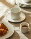 White ceramic dinnerware set by Alessi, with cups, saucers, and plates on a textured linen cloth with strong shadows.