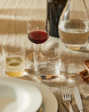 Clear glassware set by Alessi, displayed on a sunlit linen tablecloth with red wine and water.