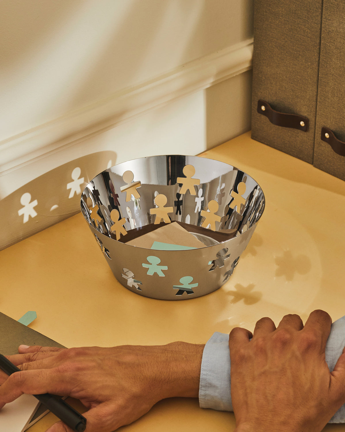 Silver metallic basket with human figure cutouts by Alessi, containing paper on a warm-toned desk.