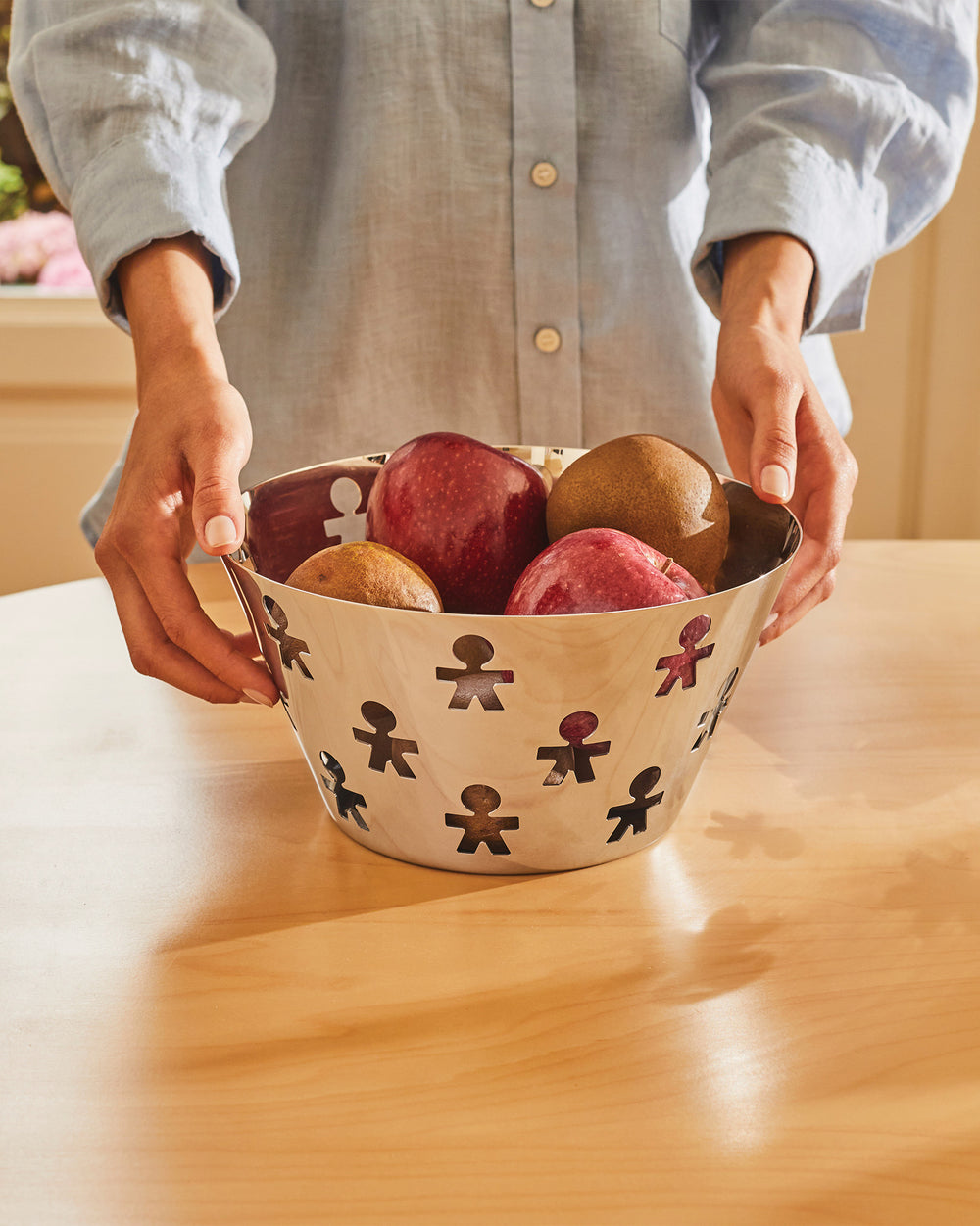 Polished metal fruit bowl by Alessi, featuring a cut-out 'Girotondo' human figure pattern, filled with fruit.