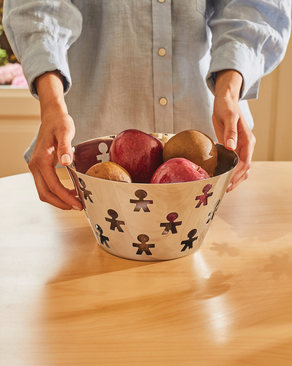 Polished metal fruit bowl by Alessi, featuring a cut-out 'Girotondo' human figure pattern, filled with fruit.