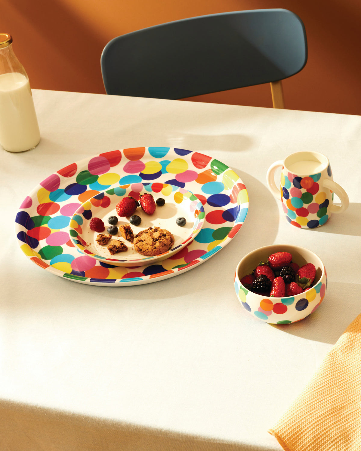Colorful polka dot oval plates, mug, and bowl by Alessi, arranged on a white tablecloth.