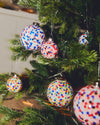 Spherical white Christmas ornament with colorful confetti dots by Alessi, hanging on a green Christmas tree.