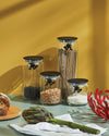 Clear glass storage jars with black lids featuring 'Gianni' a little man holding on tight by Alessi, filled with dry goods.