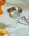 Silver-toned and clear glass ice cream cup by Alessi, with a heart-shaped spoon and yellow macaron.