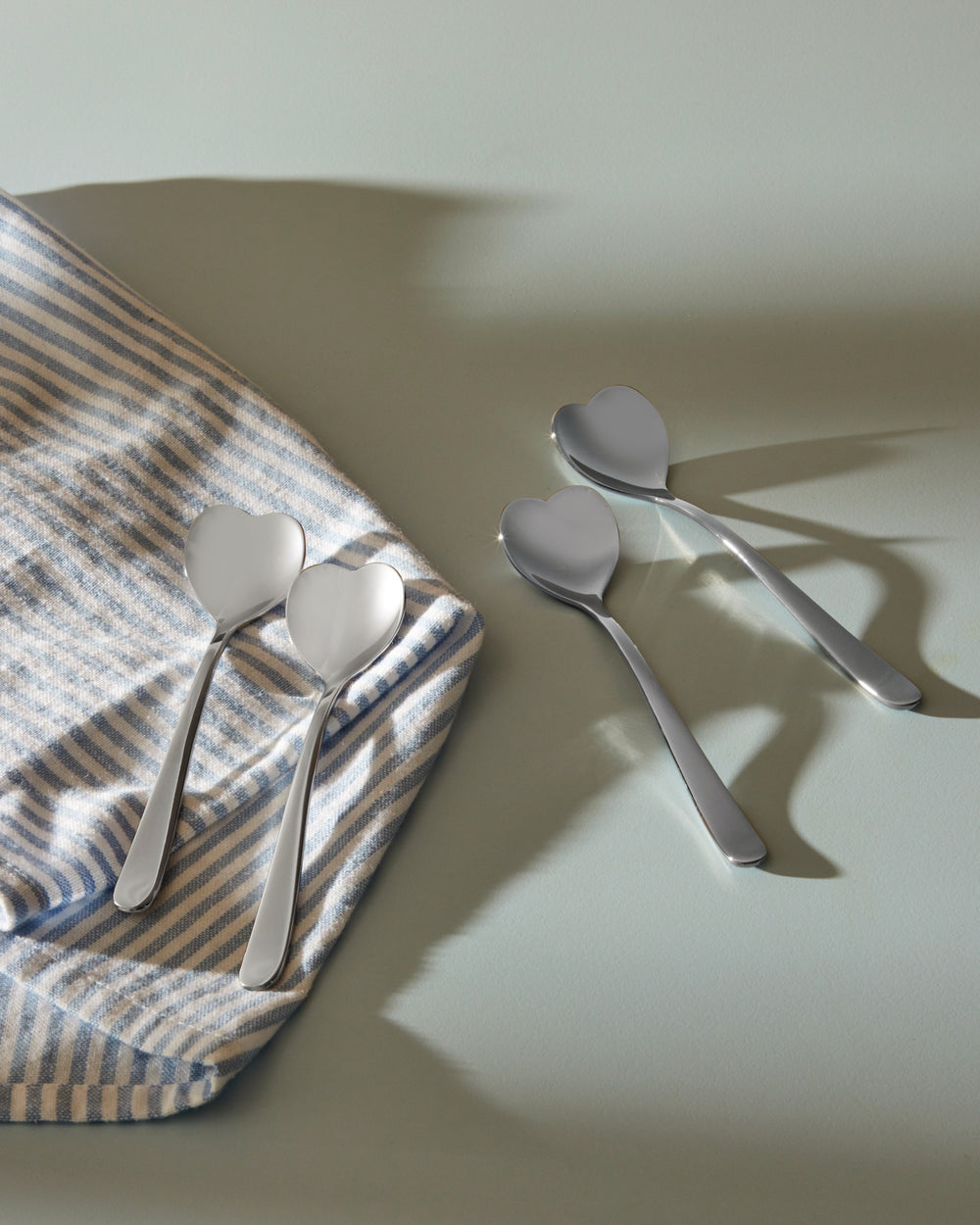 Four heart-shaped stainless steel spoons by Alessi, two on a striped napkin and two on a light green surface.