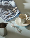 Silver heart-shaped spoons by Alessi arranged on a blue linen napkin, alongside a white cup and silver jug.