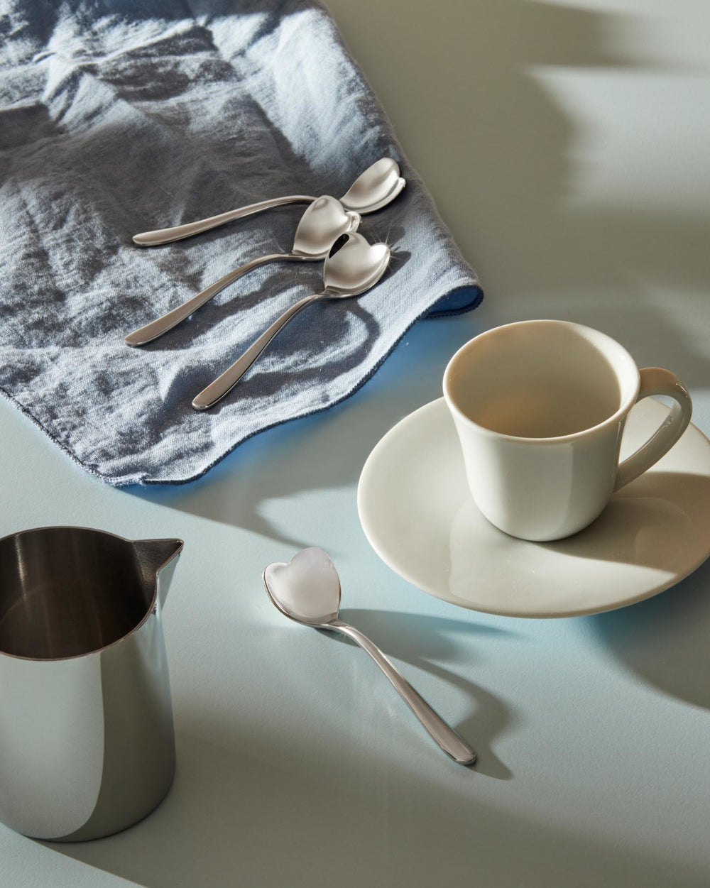 Silver heart-shaped spoons by Alessi arranged on a blue linen napkin, alongside a white cup and silver jug.