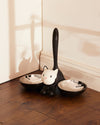 Black and white cat-shaped pet feeder with two silver bowls by Alessi, on a wooden floor.