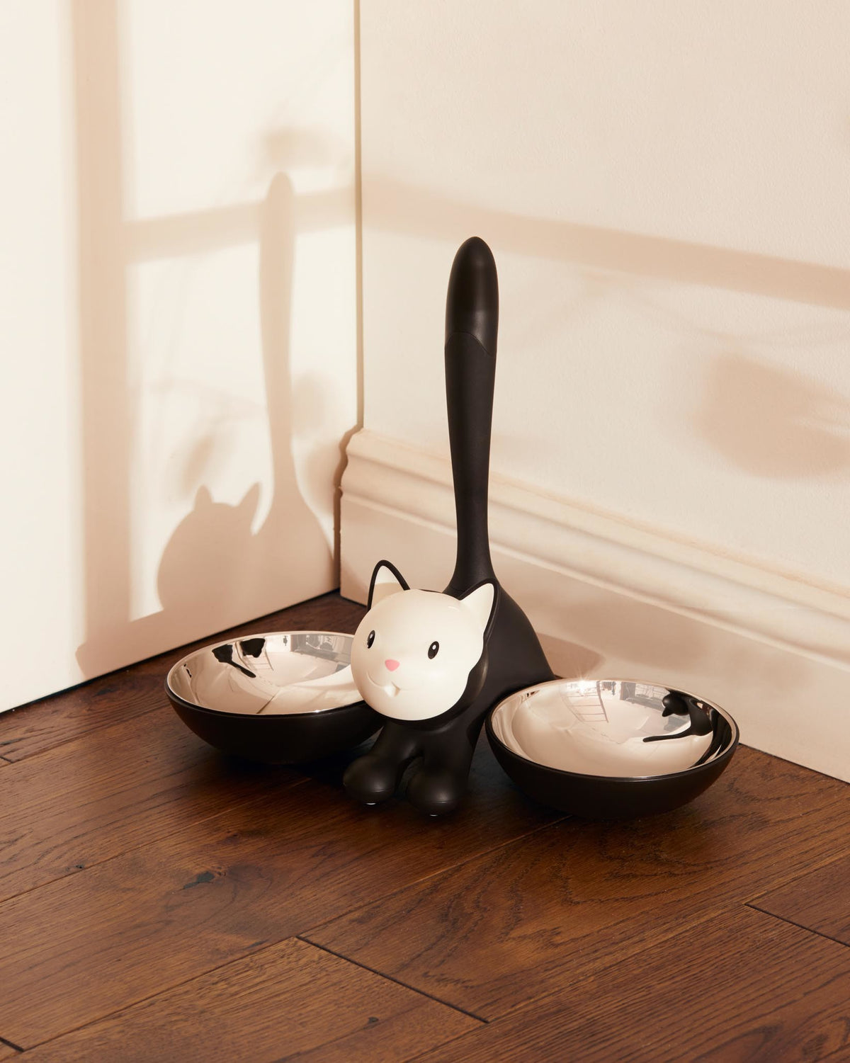 Black and white cat-shaped pet feeder with two silver bowls by Alessi, on a wooden floor.