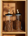 Two clear glass LulàJars by Alessi, with black lids topped by white dog and cat figurines, filled with pet food on a wooden shelf.