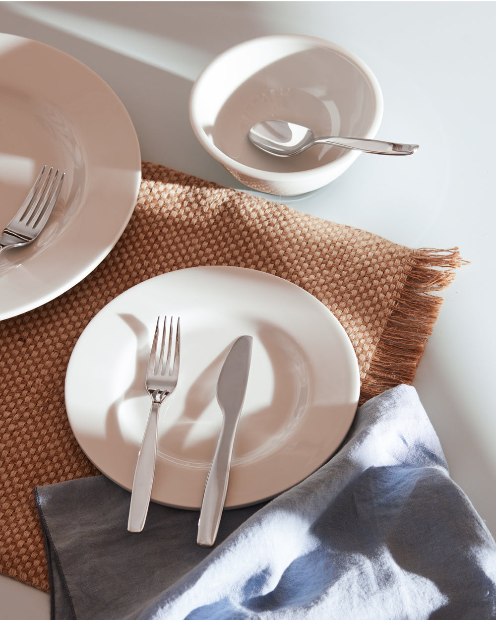 Itsumo stainless steel cutlery set by Alessi, arranged on white dinnerware with a woven placemat and blue napkin.