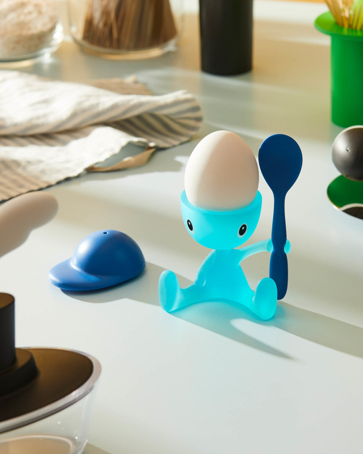 Blue translucent character egg cup by Alessi holding a dark blue spoon, with a white egg and a matching blue cap.