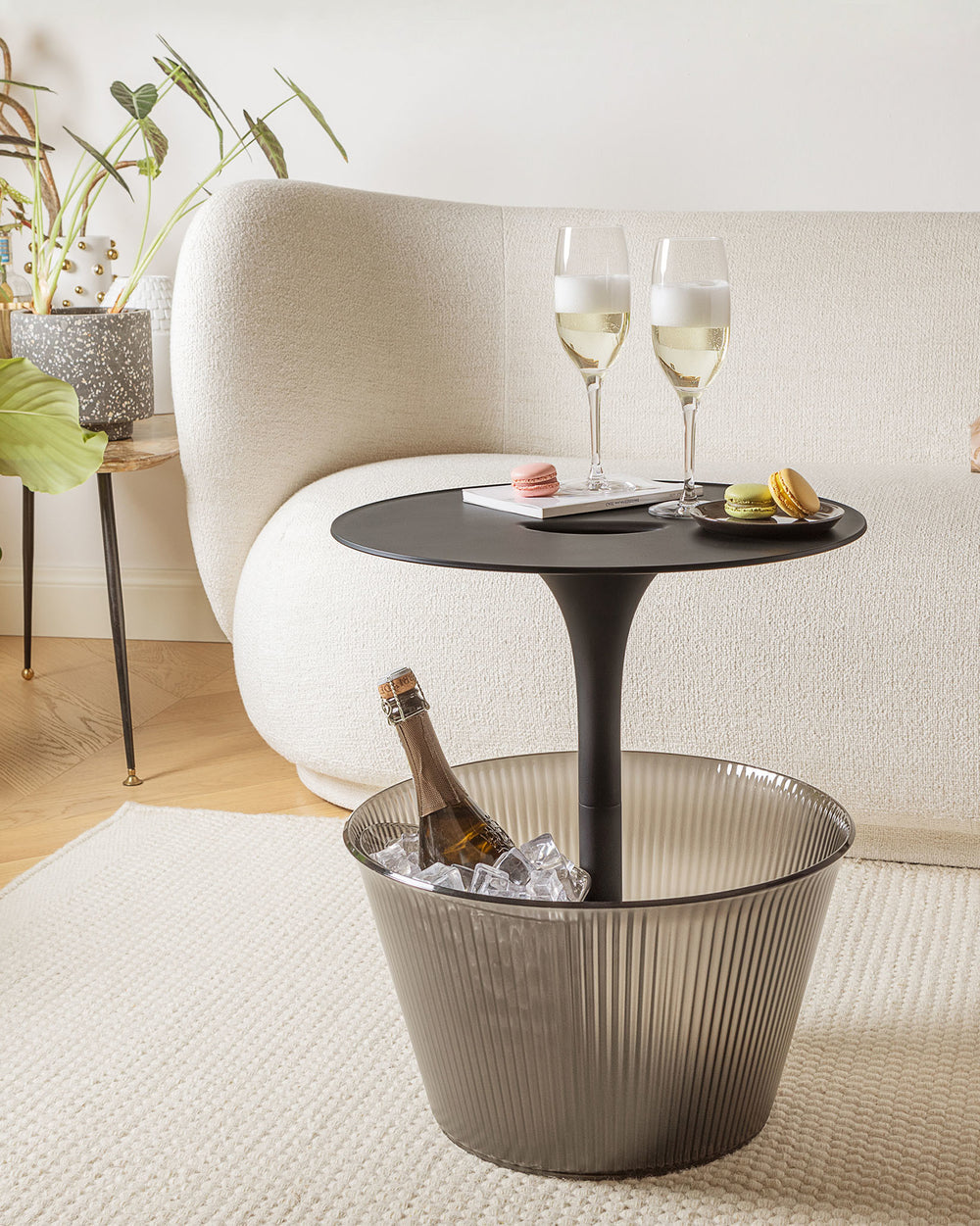 Black pedestal drinks table with smoky ribbed plastic cooler by Alessi, holding champagne and ice, with glasses and macarons.