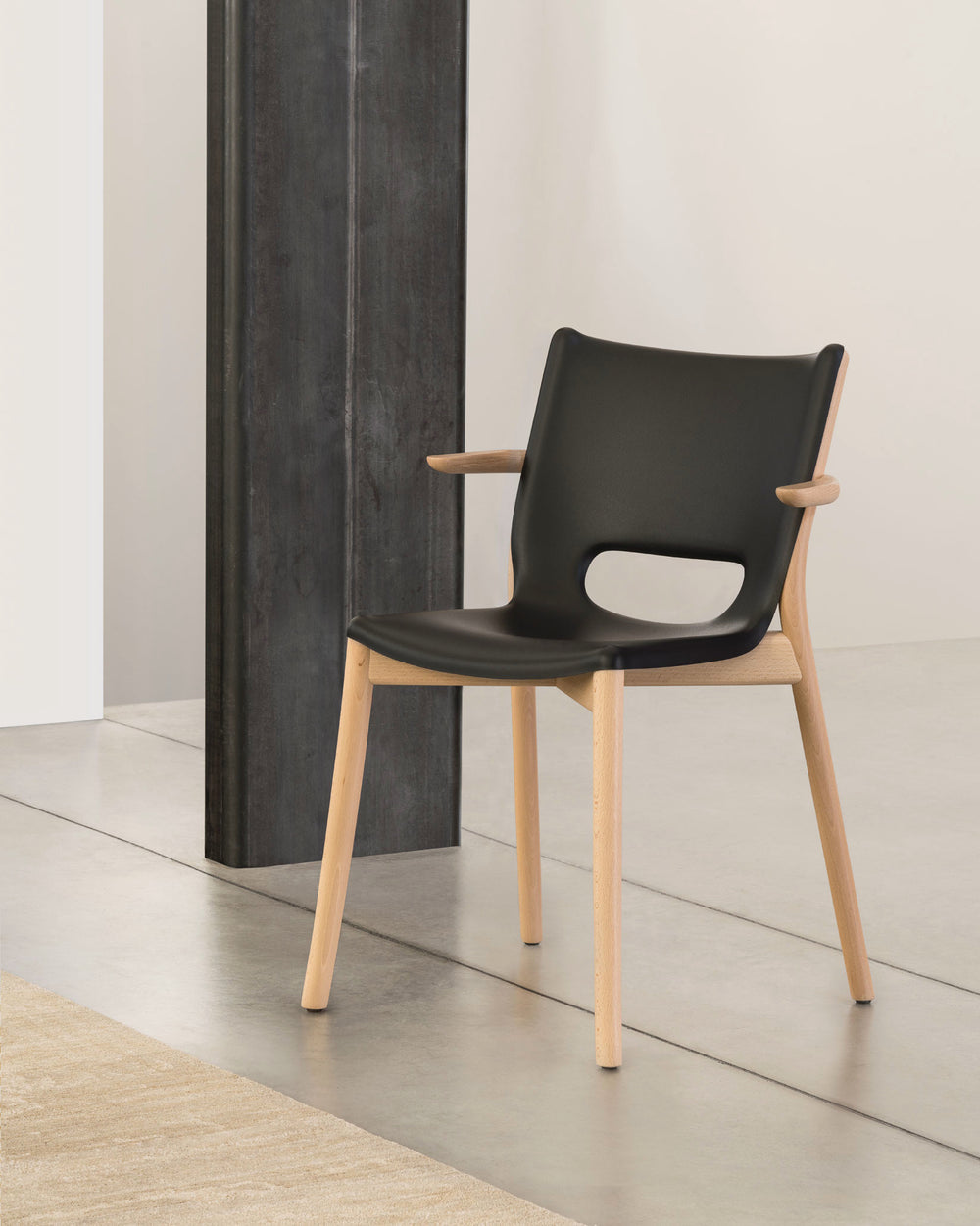 Black molded chair by Alessi with a natural wood frame, short armrests, and cutout back, on a concrete floor with a beige rug.