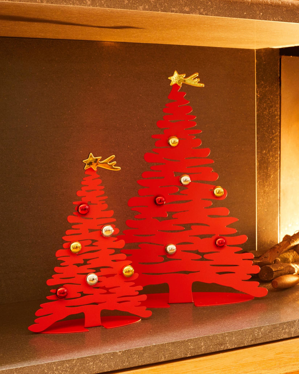 Two red metal Christmas tree decorations by Alessi, abstractly shaped, adorned with metallic baubles and a golden star.
