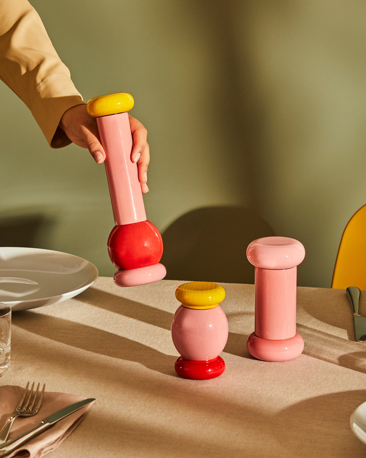 A pink, red, and yellow lacquered wood grinder by Alessi, held in a hand over a table with two matching grinders.