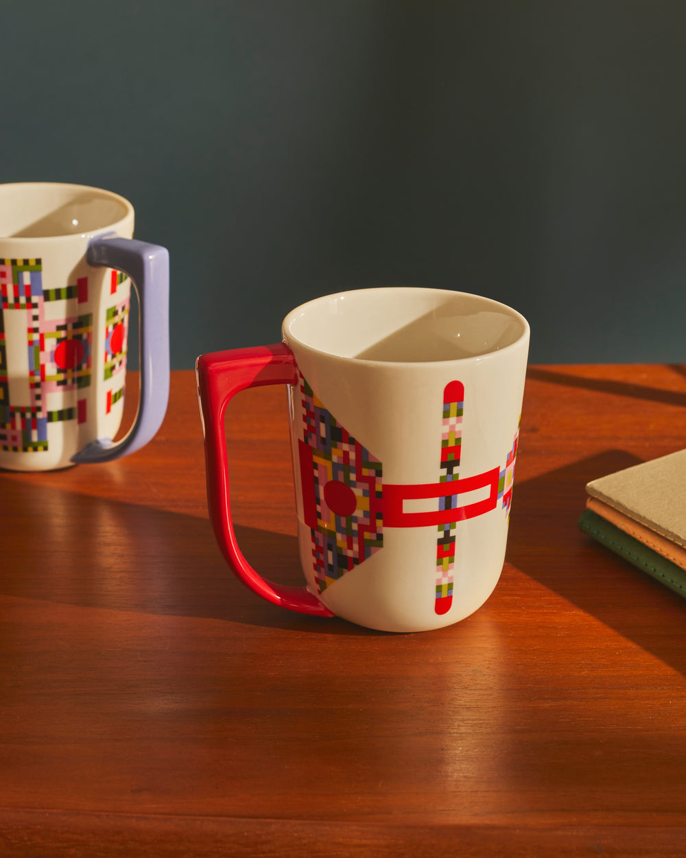 White ceramic mug by Alessi with a red handle and colorful geometric pixelated pattern. Another similar mug with a purple handle sits on a wooden table.
