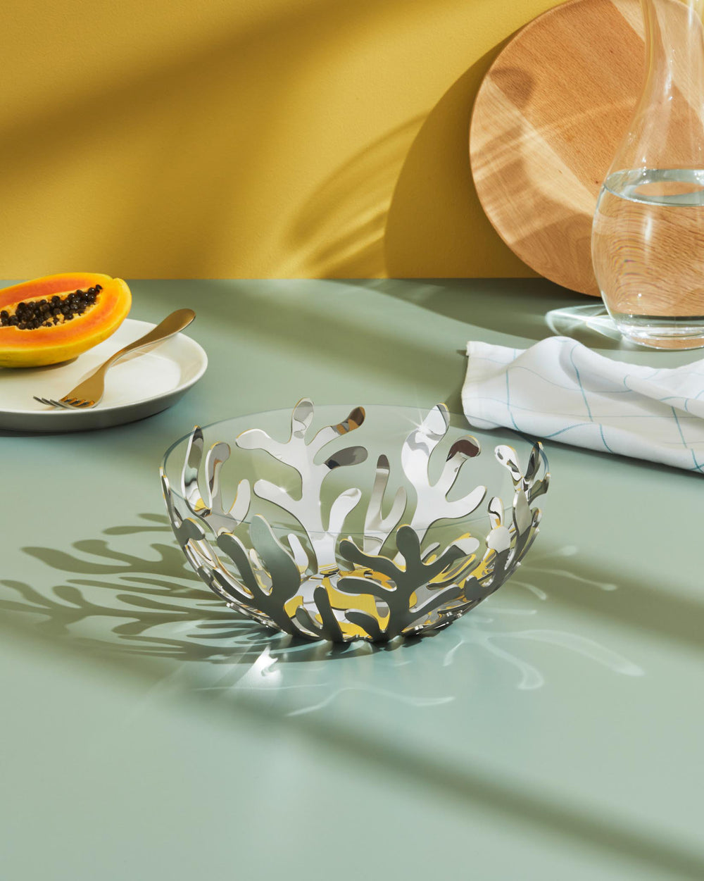 Silver metal and glass fruit bowl by Alessi, featuring an intricate coral-like cut-out design, on a light green surface.