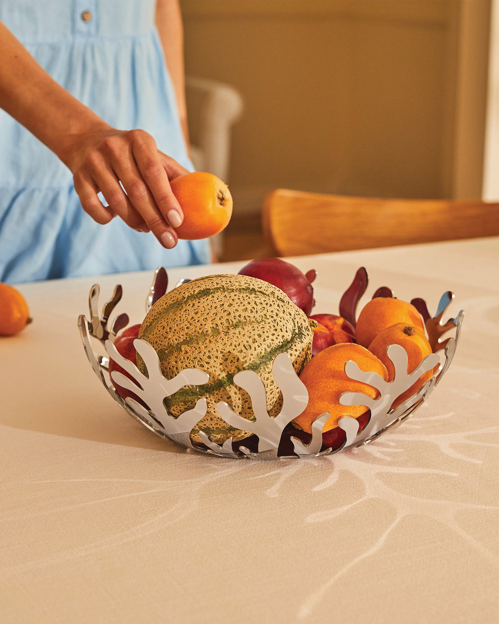 Stainless steel fruit bowl by Alessi, featuring an intricate coral design, being filled with colorful fruits by a hand in a blue dress.