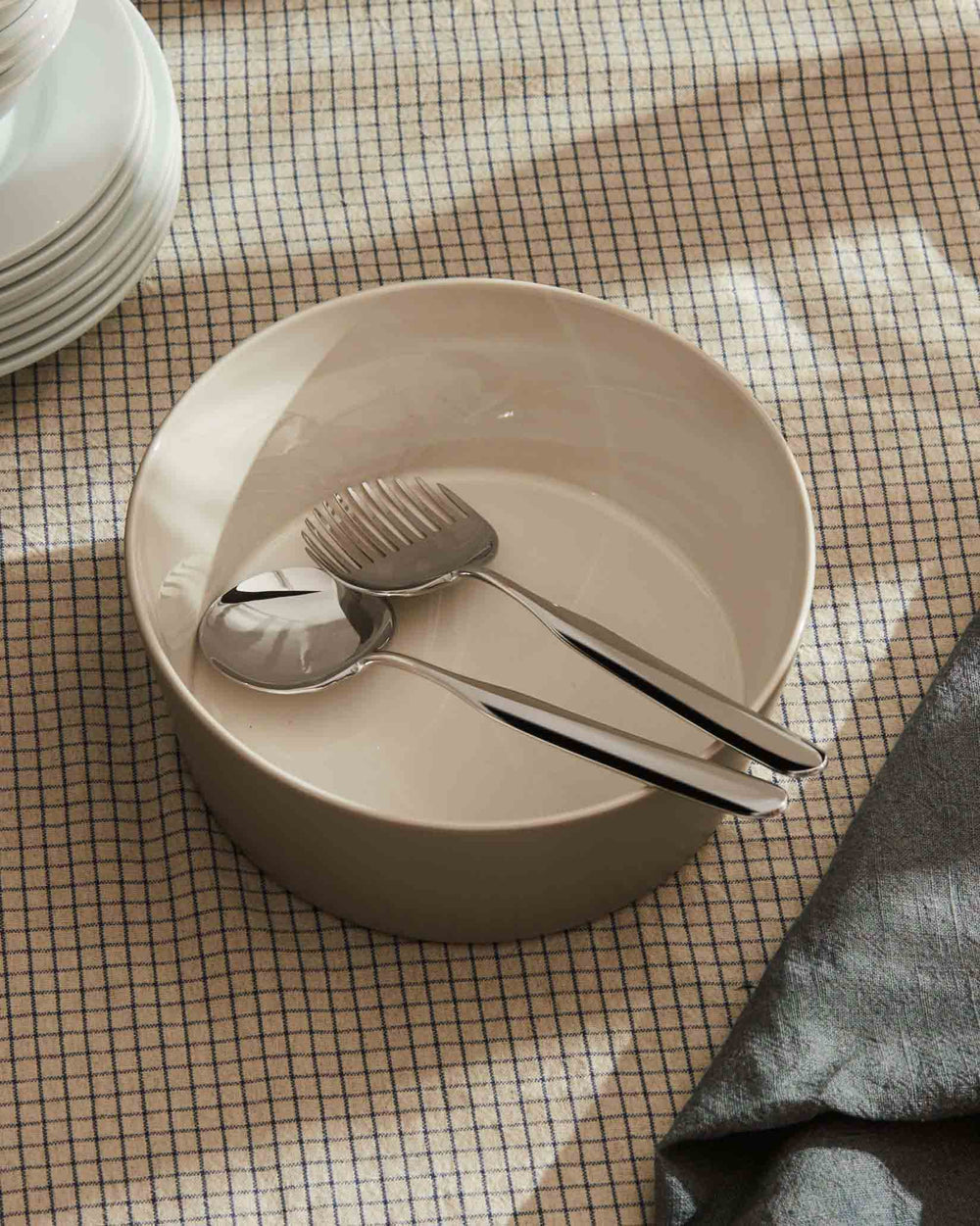 Silver serving utensils by Alessi, resting in a light ceramic bowl on a checkered tablecloth.