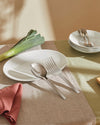 Silver metal serving cutlery by Alessi, a fork and spoon on a white oval plate with a leek and napkin.