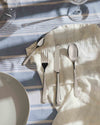 Polished silver cutlery set by Alessi, laid on a white napkin and striped blue tablecloth with glasses.
