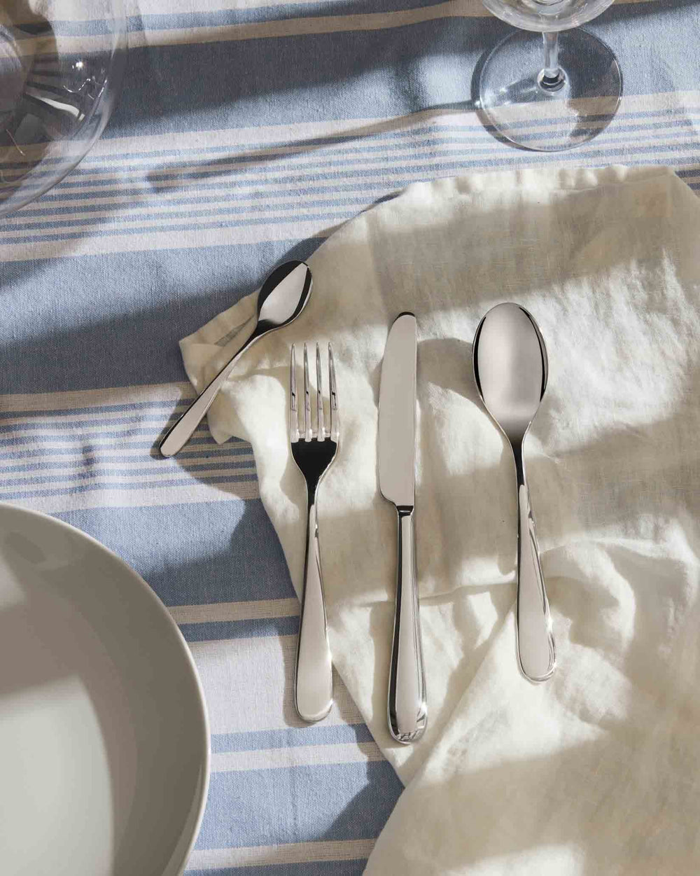 Polished silver cutlery set by Alessi, laid on a white napkin and striped blue tablecloth with glasses.