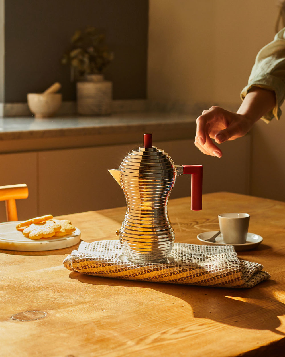 Silver ribbed coffee maker with red handle by Alessi, steaming on a wooden table with cookies and a cup.