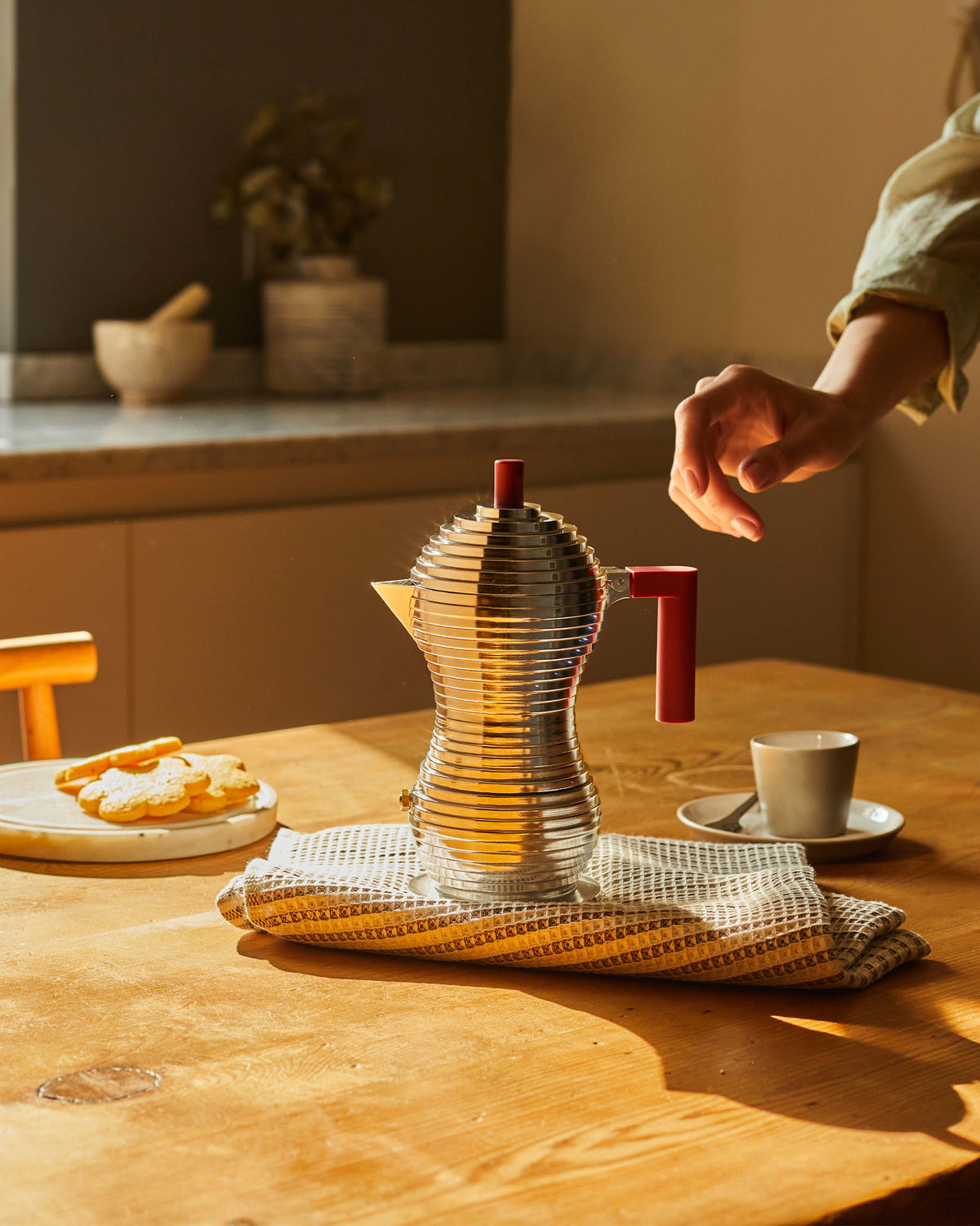 Silver ribbed coffee maker with red handle by Alessi, steaming on a wooden table with cookies and a cup.