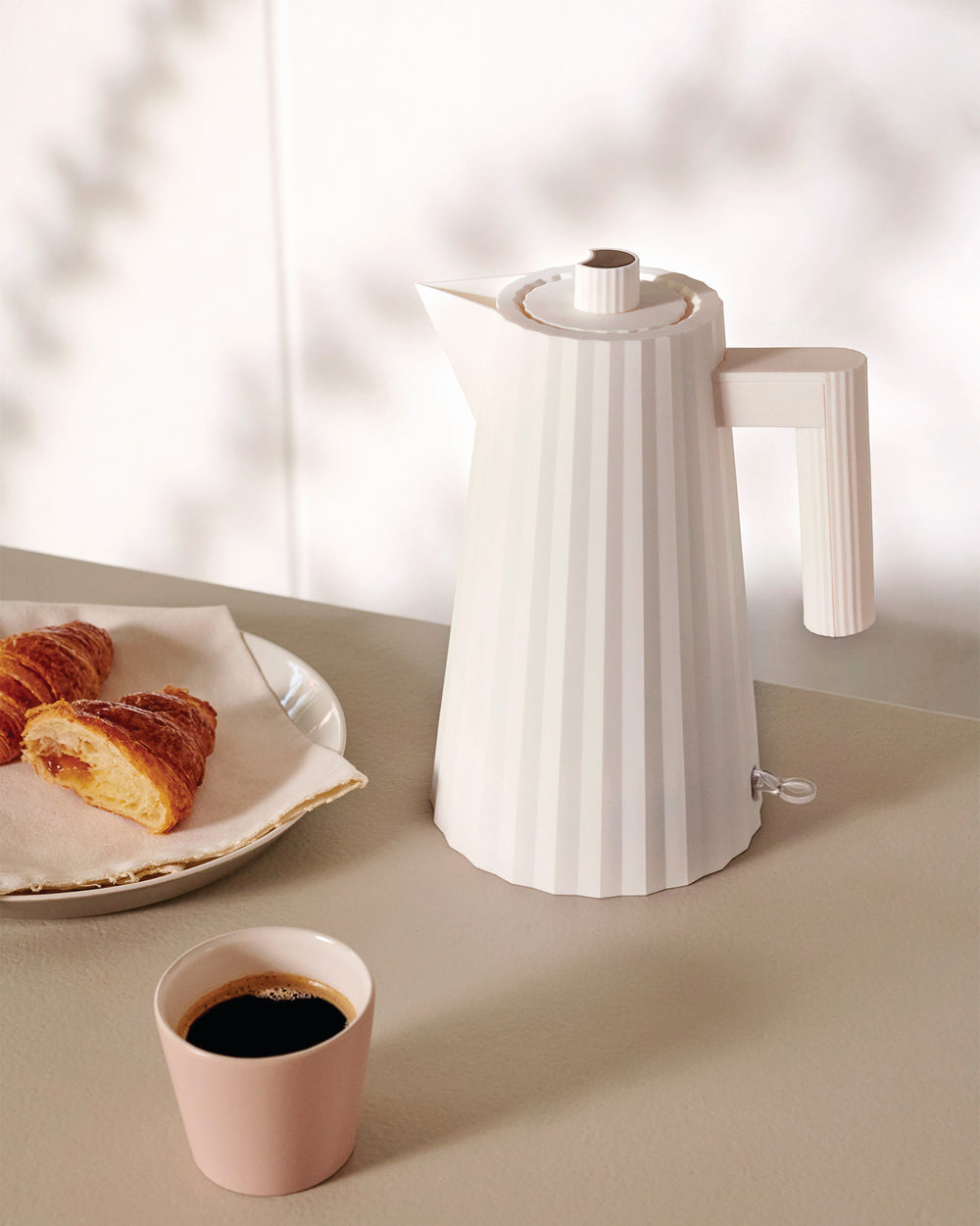 White ribbed electric kettle by Alessi, displayed on a beige counter next to a pink cup of coffee and croissants.