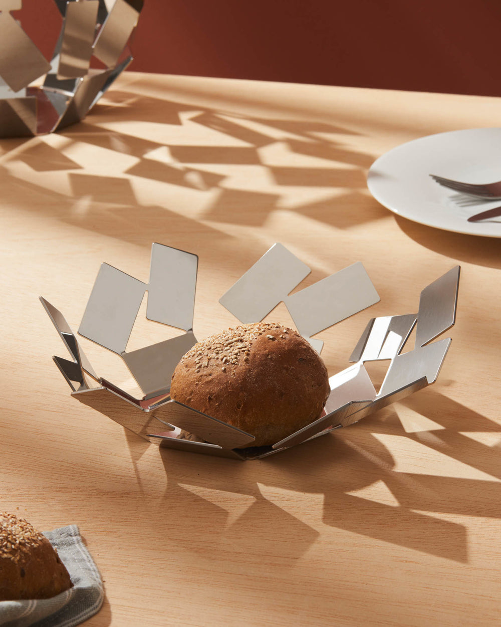 Geometric polished stainless steel bread basket by Alessi, holding a bread roll on a light wood table with geometric shadows.