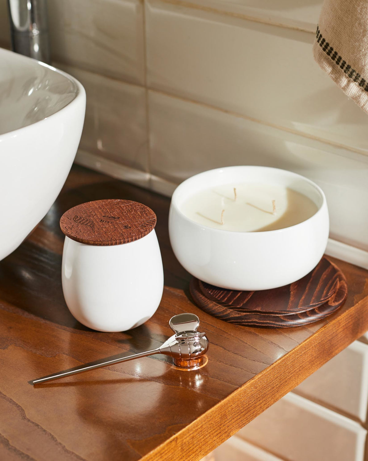 White ceramic jar by Alessi with an engraved round wooden lid, next to a multi-wick candle and shiny metal tool on a wooden surface.