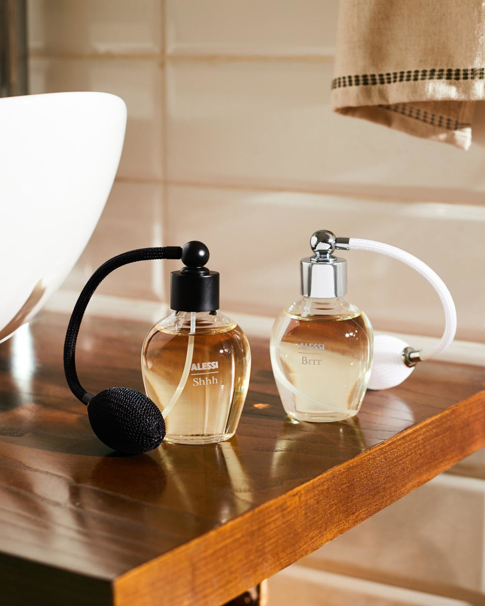Two clear glass room spray bottles by Alessi, one black with 'Shhh', one chrome and white with 'Brrr', on a wooden counter.