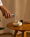 Clear diffuser refill bottle by Alessi, with blue and white floral patterns, pouring into a white ceramic diffuser vase.