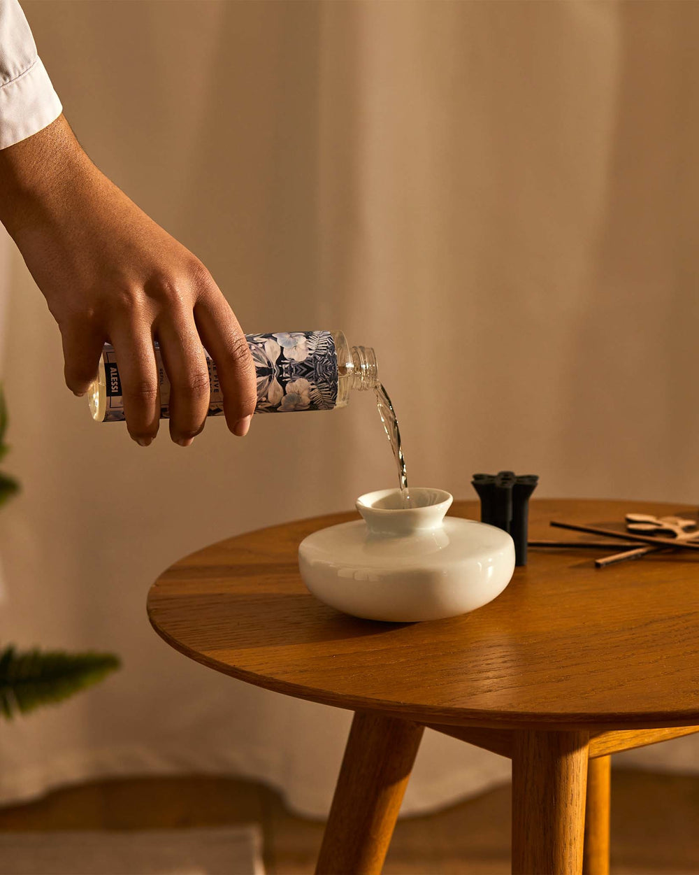 Clear diffuser refill bottle by Alessi, with blue and white floral patterns, pouring into a white ceramic diffuser vase.