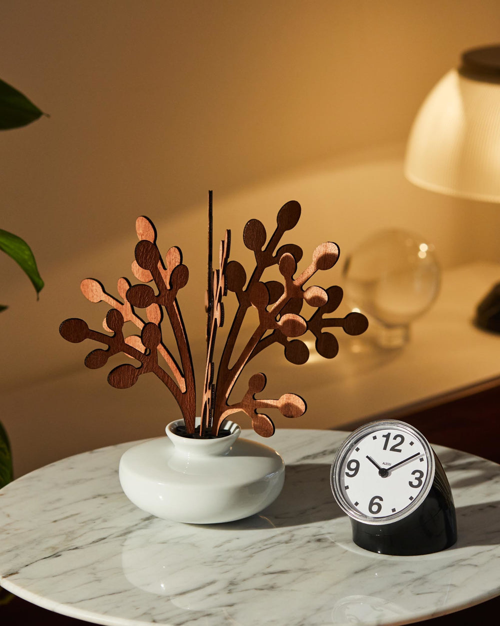 Laser-cut wooden room diffuser by Alessi, featuring branching elements in a white vase, beside an analog clock on a marble table.