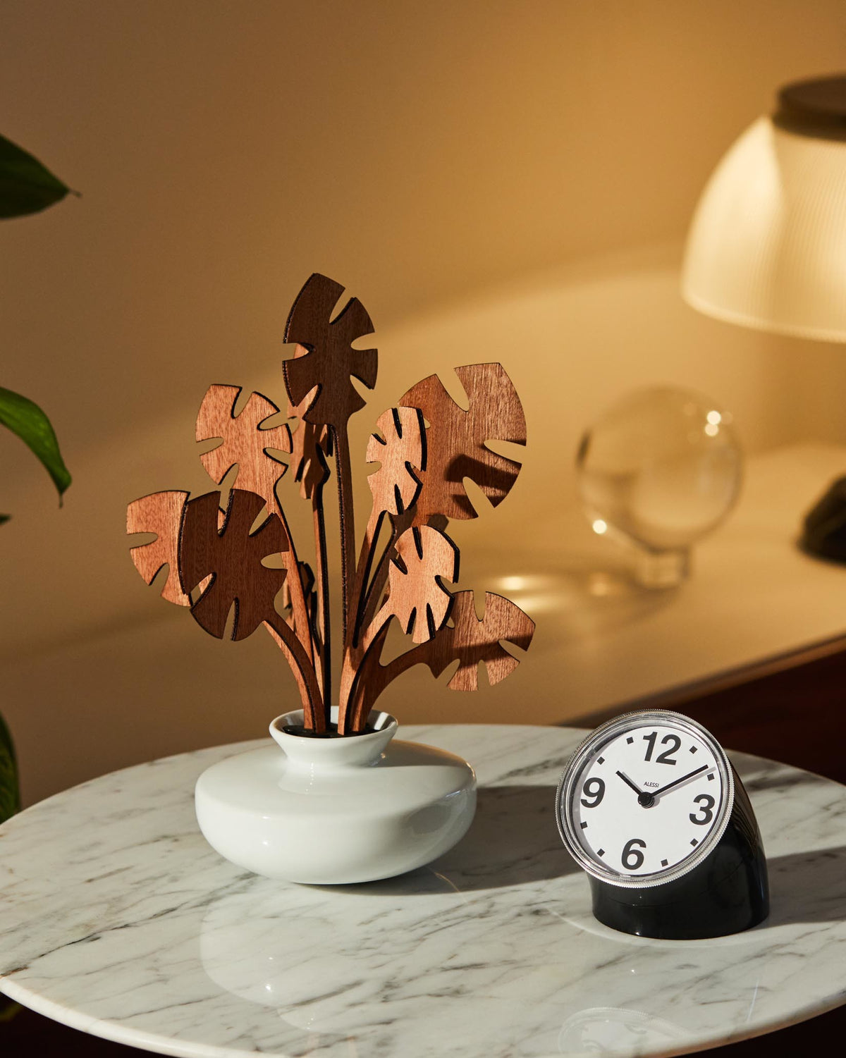 A decorative wooden plant sculpture by Alessi, featuring multiple dark brown leaves in a white vase on a marble table.