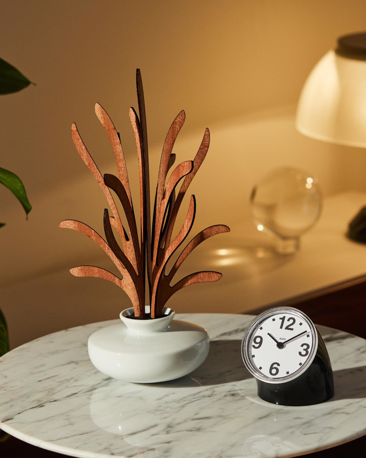 Wooden diffuser in a white ceramic vase by Alessi, arranged on a marble table with a small clock.