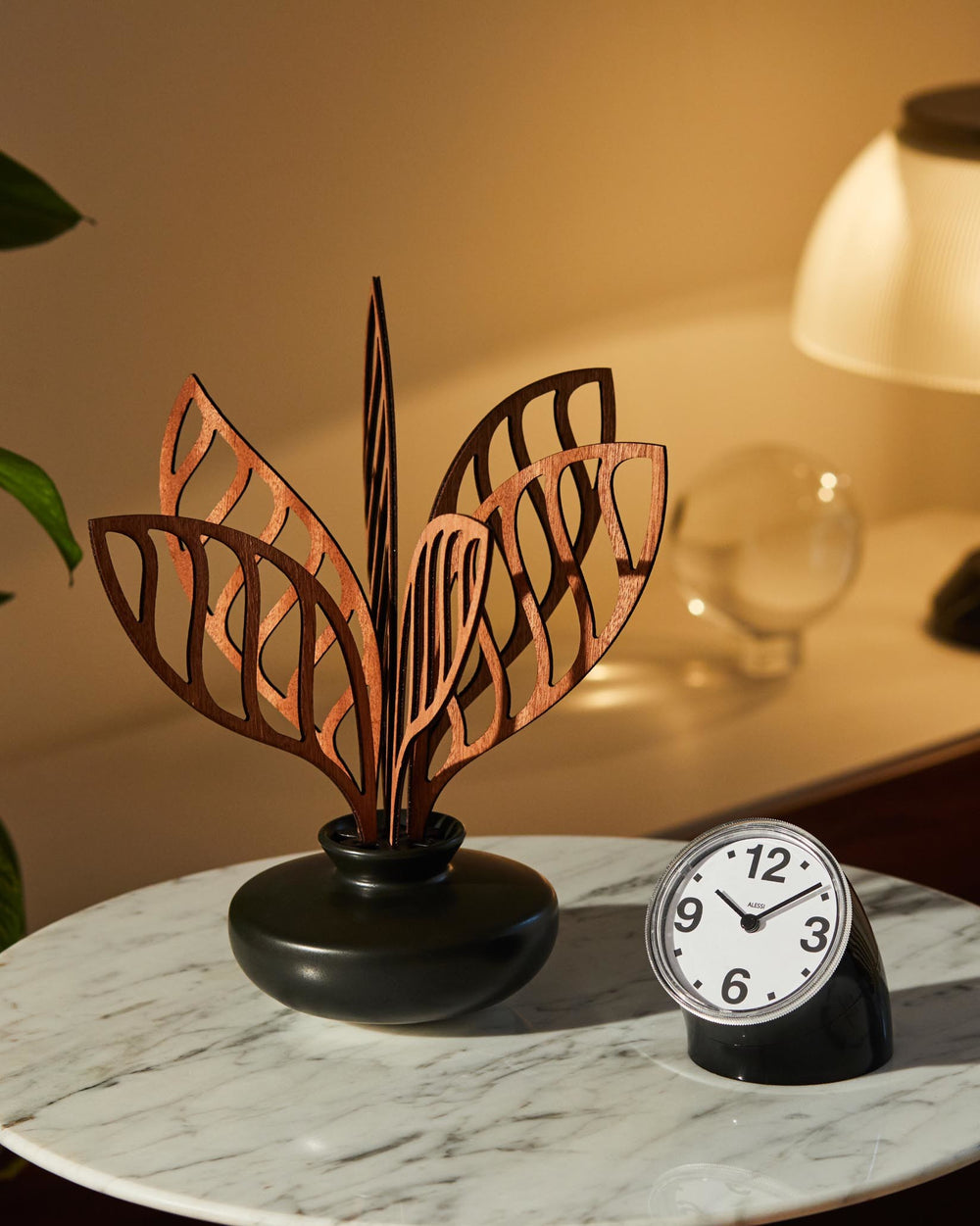 Decorative diffuser by Alessi with dark brown wooden leaf-shaped elements in a black matte vase, on a marble table with a small clock.